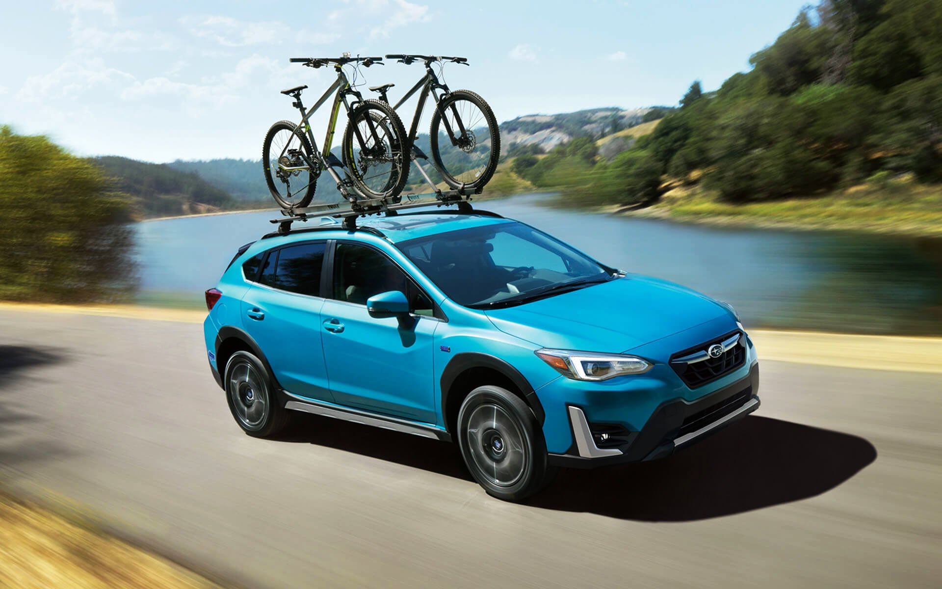 A blue Crosstrek Hybrid with two bicycles on its roof rack driving beside a river | Neil Huffman Subaru in Louisville KY